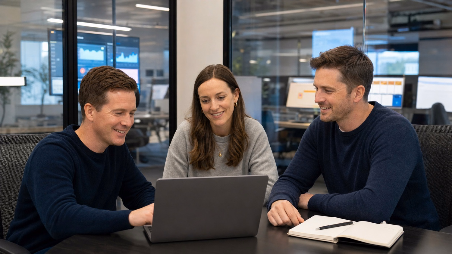Three people are seated around a small, glass-walled conference room table, disc